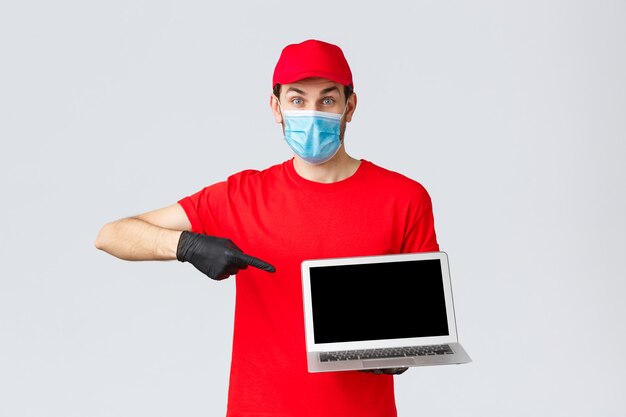 고객 지원, covid-19 배달 패키지, 온라인 주문 처리 개념. 빨간색 유니폼, 장갑, 코로나바이러스의 얼굴 마스크를 쓴 열정적인 택배, 노트북 화면을 가리키는