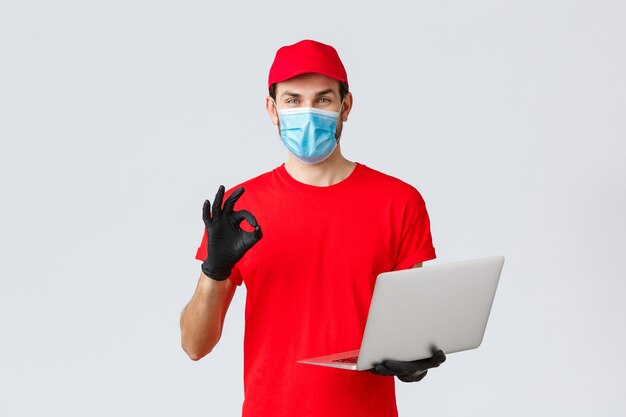 Customer support, covid-19 delivery packages, online orders processing concept. Smiling courier in face mask and gloves guarantee safety of parcel, processing order, show okay sign, hold laptop
