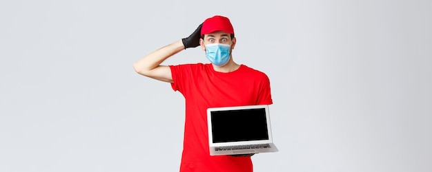 Поддержка клиентов covid19 доставка пакетов онлайн концепция обработки заказов Смущенный и взволнованный курьер в красной униформе маска для лица и перчатки выглядят нервными, показывают экран ноутбука