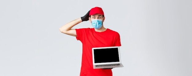 Customer support covid19 delivery packages online orders processing concept Confused and worried courier in red uniform face mask and gloves look nervous show laptop screen