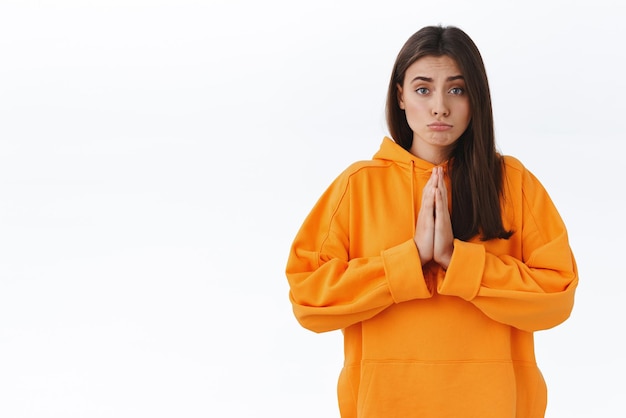 Cute adorable brunette female begging for your help lend money press hands together in pray make hopeful lovely expression as pleading apologizing saying please stand white background