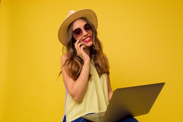 Cute adorable woman with dark wavy hair is smiling with happy emotions she is talking on the phone and holding laptop over yellow background