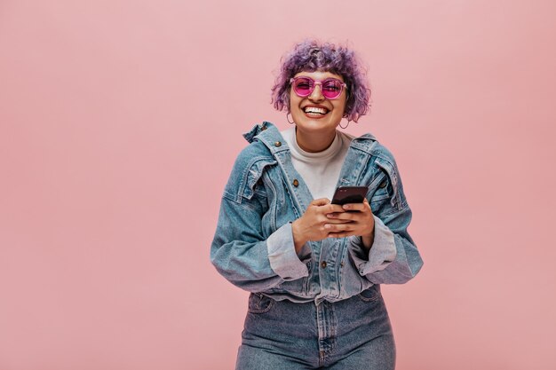 Cute adult lady with bright haircut in sunglasses laughs. Smiling woman with curly hair holding phone.