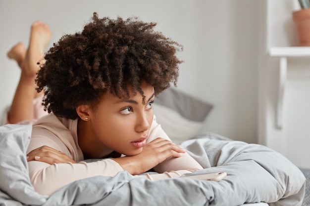 Cute African girl lies in bed, leans on hands, has thoughtful expression, enjoys comfort