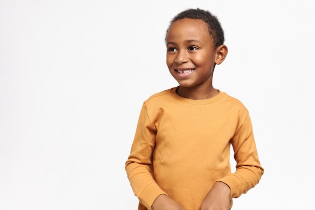 Cute Afro American schoolboy in yellow sweatshirt posing against white wall with copy space for your information