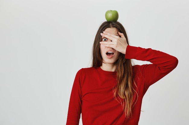 Cute ambushed girl feel scared of her life, cover eyes, peeking through fingers as hold green apple target on head