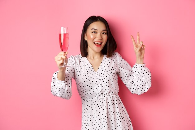 Cute asian female model drinking champagne celebrating on party and showing peace sign smiling happy...