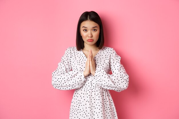 Cute asian girl asking for help, begging for favour and looking innocent at camera, pleading against pink background