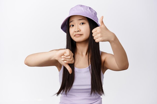 Cute asian girl asking your opinion showing thumbs up and down and looking at camera need advice standing over white background