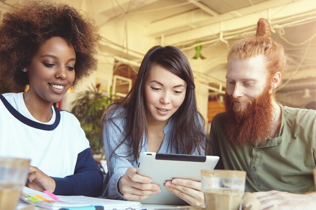 Cute Asian girl in blue shirt holding tablet showing presentation to partners who looking at screen with cheerful expression.