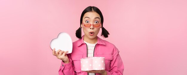 Cute asian girl giving you gift in heart shaped box kissing and smiling concept of holiday and celebration standing over pink background