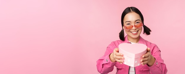 ピンクの背景の上に立っている休日とお祝いの概念にキスと笑顔のハート型のボックスであなたに贈り物を与えるかわいいアジアの女の子