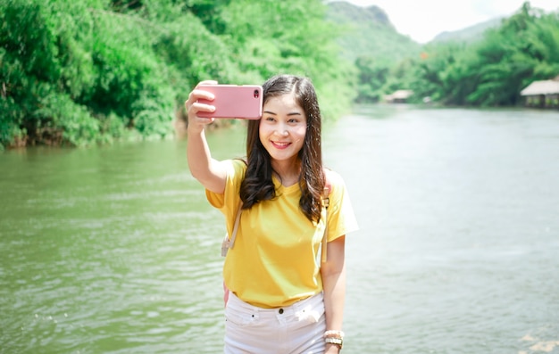 귀여운 아시아 소녀, 노란색 티셔츠와 분홍색 배낭에 그녀의 여행에서 그녀는 셀카를 찍고 녹색 자연 장소와 많은 순간에 포즈를 취했다. 프리미엄 사진