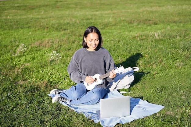 Cute asian girl musician studying looks at tutorial on her laptop online tutor shows how to play ukulele student sits on grass in park on sunny day