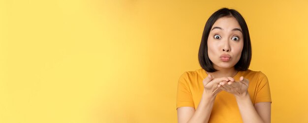 Cute asian girl sending air kiss blowing mwah with puckered lips standing over yellow background