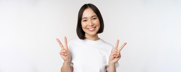 Симпатичная азиатская девушка, демонстрирующая мир, улыбается и выглядит счастливой перед камерой в белой футболке на студийном фоне