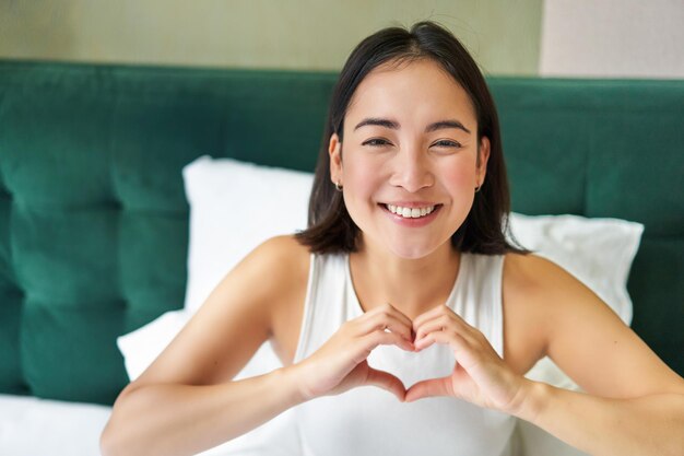 cute asian girl shows heart love sign lying in bed and smiling enjoying mornings lazy days in bedroo