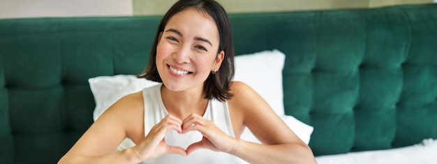 Cute asian girl shows heart love sign lying in bed and smiling enjoying mornings lazy days in