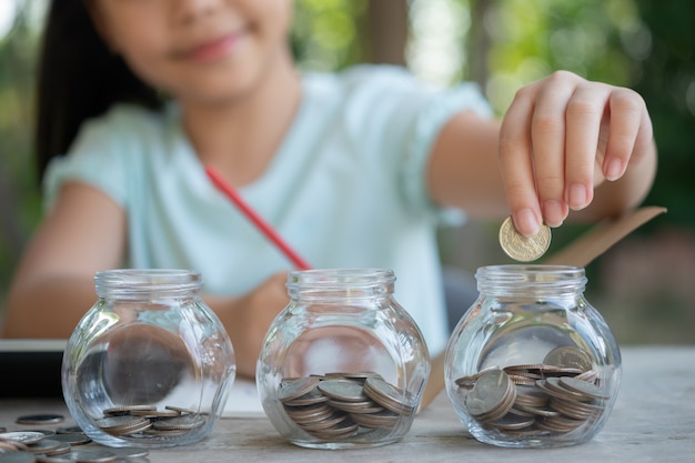 お金の山を作るコインで遊んでいるかわいいアジアの女の子、貯金箱、ガラスの瓶にお金を貯める子供。貯めたコインを数える子ども、将来のコンセプトについて学ぶ子ども。