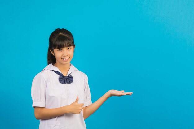Милая азиатская школьница делая большие пальцы руки вверх показывать и раскрывает вашу руку на сини.