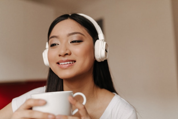 Cute Asian woman in big headphones is smiling and holding cup of tea