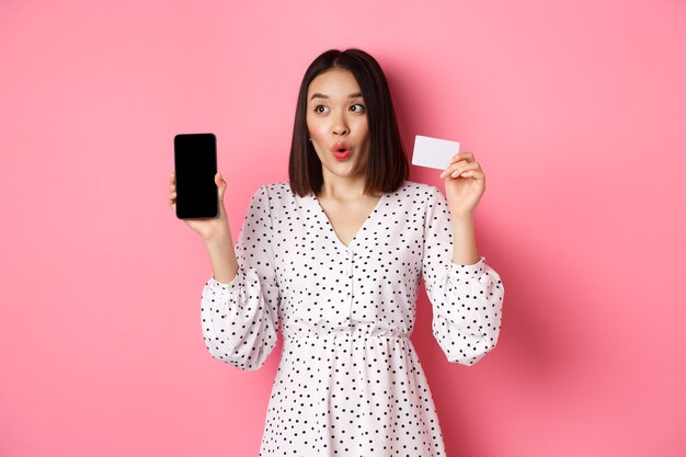 Cute asian woman shopping online showing bank credit card and mobile screen smiling and looking left...