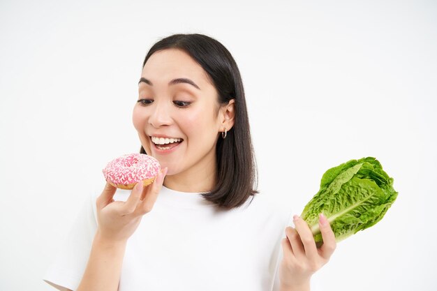 귀여운 아시아 여성은 다이어트를 건너뛰고 흰색 배경 위에 서 있는 맛있는 유약을 바른 도넛을 한 입 먹습니다