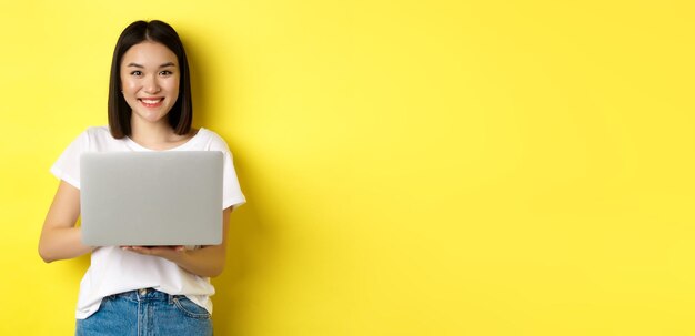 Cute asian woman studying on laptop and smiling standing in white tshirt and jeans against yellow ba