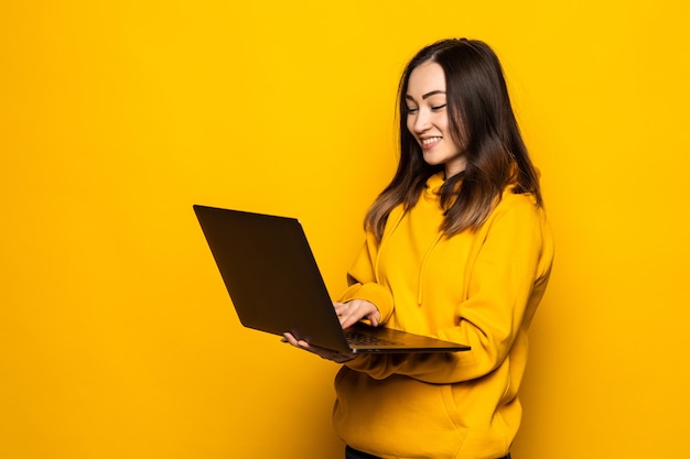 Симпатичная азиатская женщина учится на ноутбуке и улыбается, стоя у желтой стены Бесплатные Фотографии