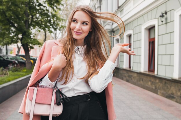 Cute attractive stylish smiling woman walking city street in pink coat spring fashion trend holding purse, elegant style