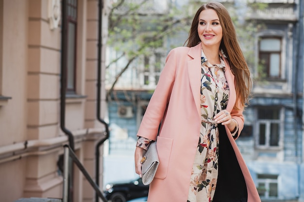 Cute attractive stylish smiling woman walking city street in pink coat spring fashion trend holding purse