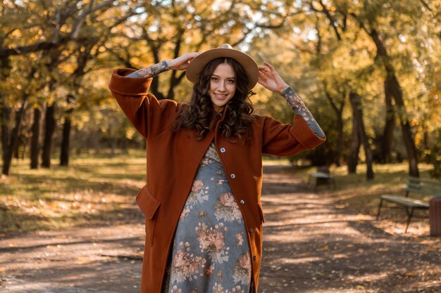Симпатичная привлекательная стильная улыбающаяся женщина с вьющимися волосами гуляет в парке, одетая в платье с принтом и теплое пальто осенняя модная мода, уличный стиль