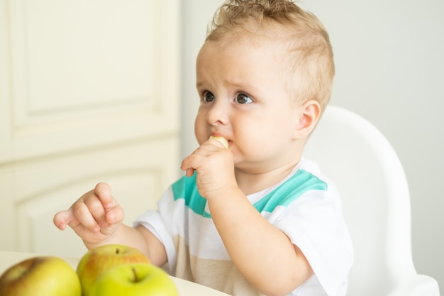 Cute baby boy sitting at the table in child chair eating apple on white kitchen