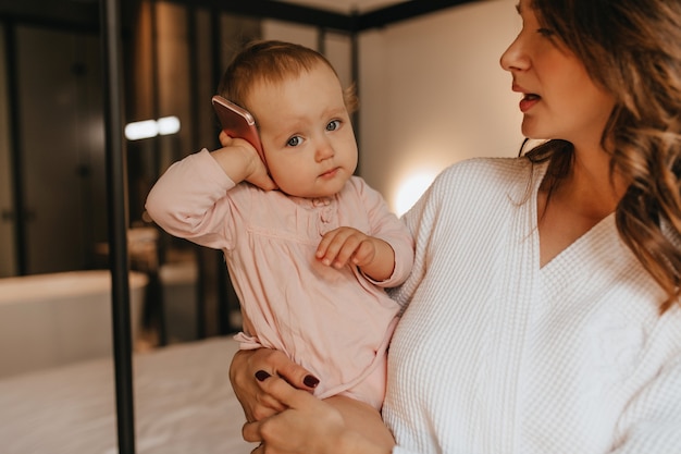 Cute baby in gently pink home clothes is holding phone while her mother hugs her on background of bed.
