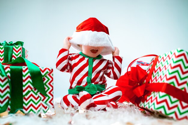 Cute baby girl 1 year old wearing santa hat posing over Christmas decorations with gifts. Sitting on floor with Christmas ball. Holiday season.