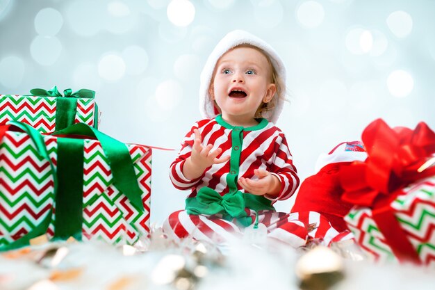 Cute baby girl 1 year old wearing santa hat posing over Christmas decorations with gifts. Sitting on floor with Christmas ball