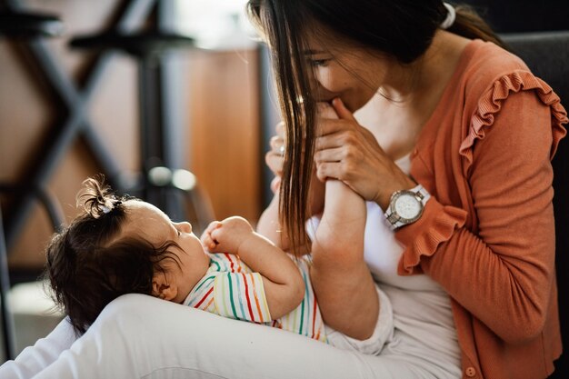 Cute baby girl lying on mother's lap while she is kissing her little toes at home