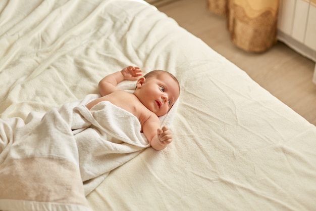 Cute baby wrapped towel after shower lying on bed on white plaid, looking away and raising hands, infant studying outward things, tiny boy or girl indoor.