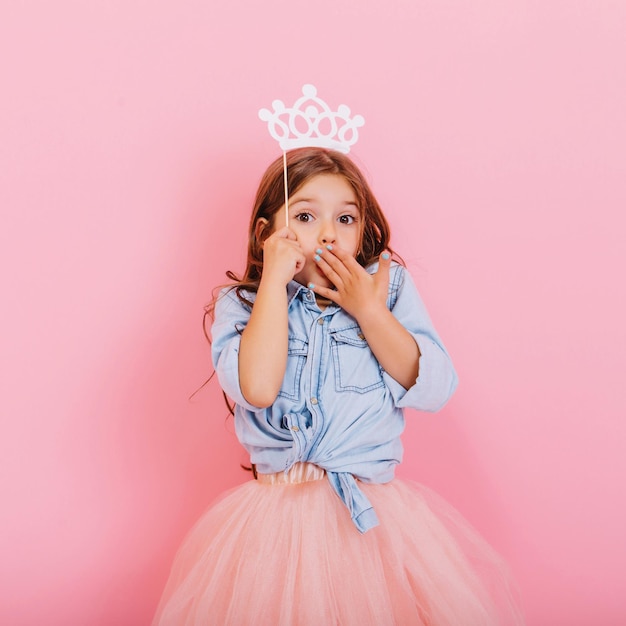 Cute beautiful carnival kid having fun isolated on pink background. Pretty little girl with long brunette hair, in tulle skirt, with white crown on head expressing wondering to camera