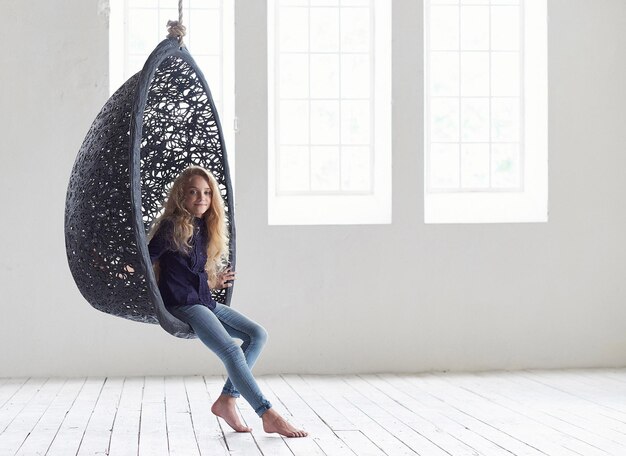 A cute beautiful little girl in casual clothes sits on a suspended chair in an empty studio.