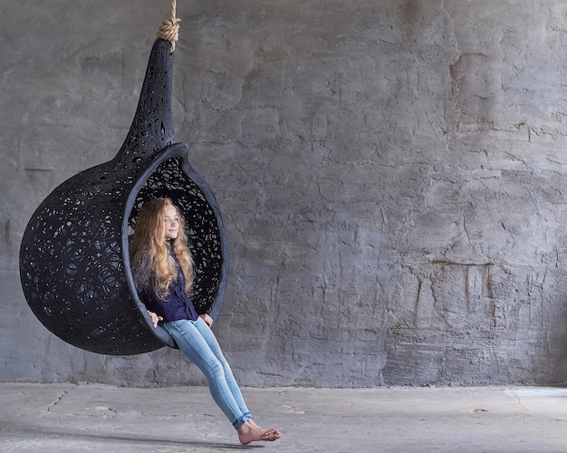 A cute beautiful little girl in casual clothes sits on a suspended chair in an empty studio.