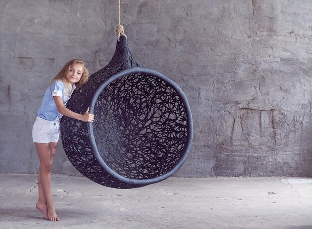 A cute beautiful little girl in casual clothes standing near a suspended chair in an empty studio.