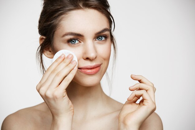 Cute beautiful natural brunette girl cleaning face with cotton sponge smiling looking at camera over white background. Cosmetology and spa.