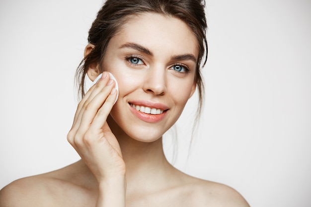 Cute beautiful natural brunette girl cleaning face with cotton sponge smiling looking at camera over white background. Cosmetology and spa.