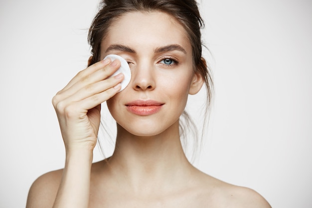 Fronte di pulizia della bella ragazza castana naturale sveglia con la spugna di cotone che sorride esaminando macchina fotografica sopra fondo bianco. cosmetologia e spa.