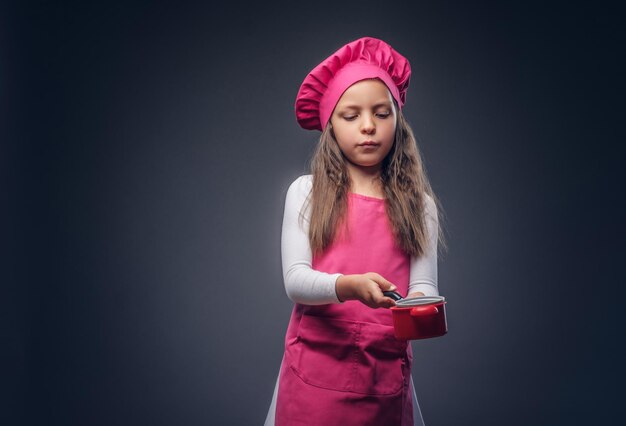 분홍색 요리복을 입은 귀여운 여학생이 스튜디오에서 조리기구를 들고 있습니다. 어두운 배경에 고립.