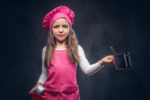 Cute beautiful schoolgirl dressed in a pink cook uniform holds cookware at a studio. Isolated on the dark textured background.