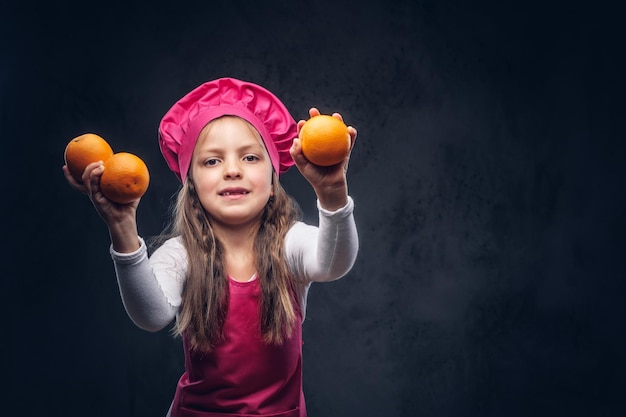 Cute beautiful schoolgirl dressed in a pink cook uniform holds oranges at a studio. Isolated on a dark textured background.