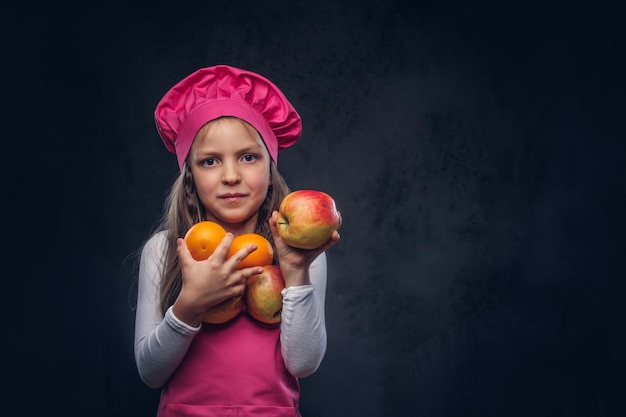 Cute beautiful schoolgirl dressed in a pink cook uniform holds oranges at a studio. Isolated on a dark textured background.