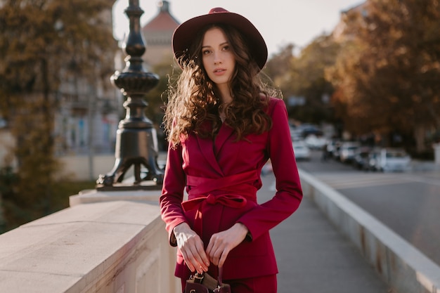 Cute beautiful stylish woman in purple suit walking in city street, spring summer autumn season fashion trend wearing hat, holding purse